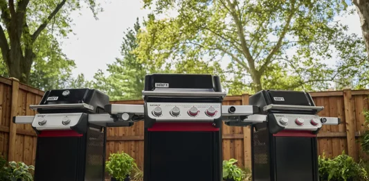 Three new 2025 Weber Spirit models side-by-side on a patio in a backyard on a warm spring day. From left to right is a two-burner, a four-burner, and three-burner 2025 Weber Spirit models