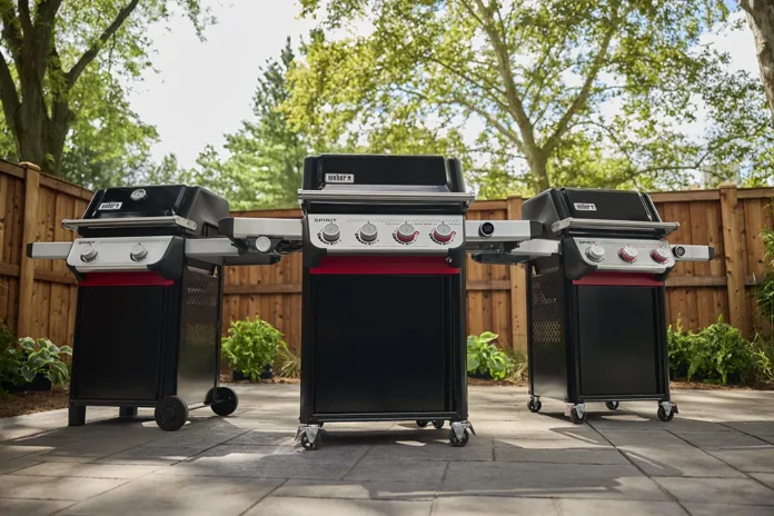 Three new 2025 Weber Spirit models side-by-side on a patio in a backyard on a warm spring day. From left to right is a two-burner, a four-burner, and three-burner 2025 Weber Spirit models